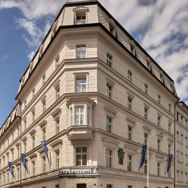 Großes weißes Gebäude mit vielen Fenstern, Wolkenhimmel und blauen Flaggen mit dem Namen „Falkensteinsteiner“.