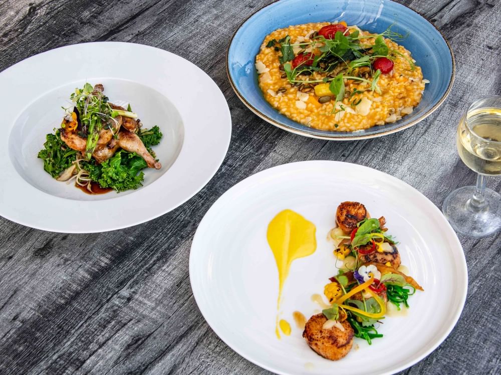 Three plates of food with various dishes and a wine glass on a wooden table.
