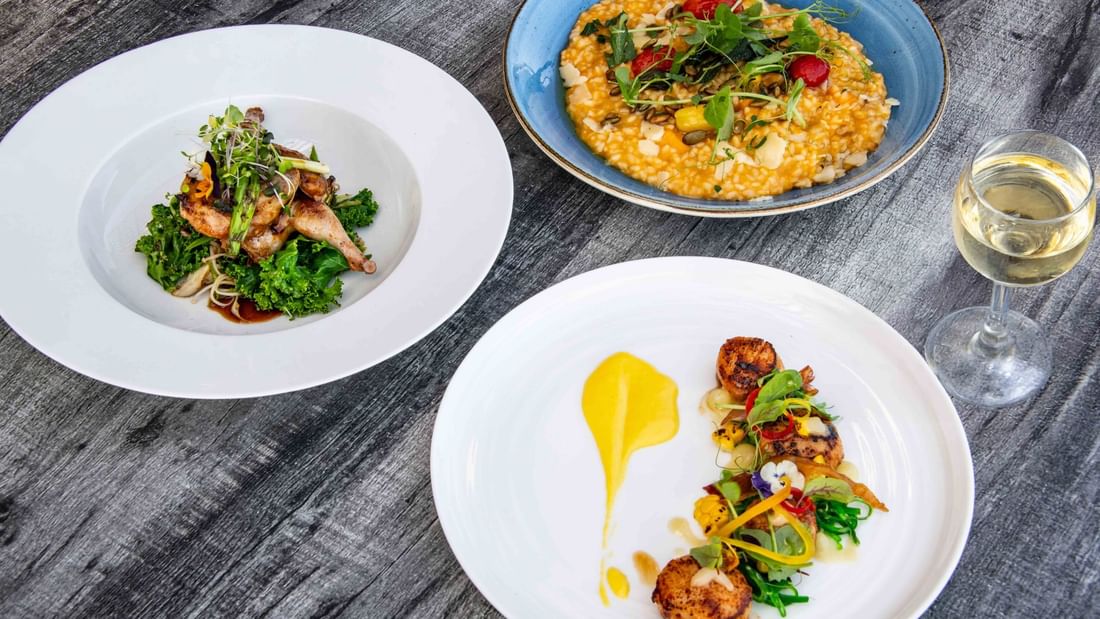 Three plates of food with various dishes and a wine glass on a wooden table.