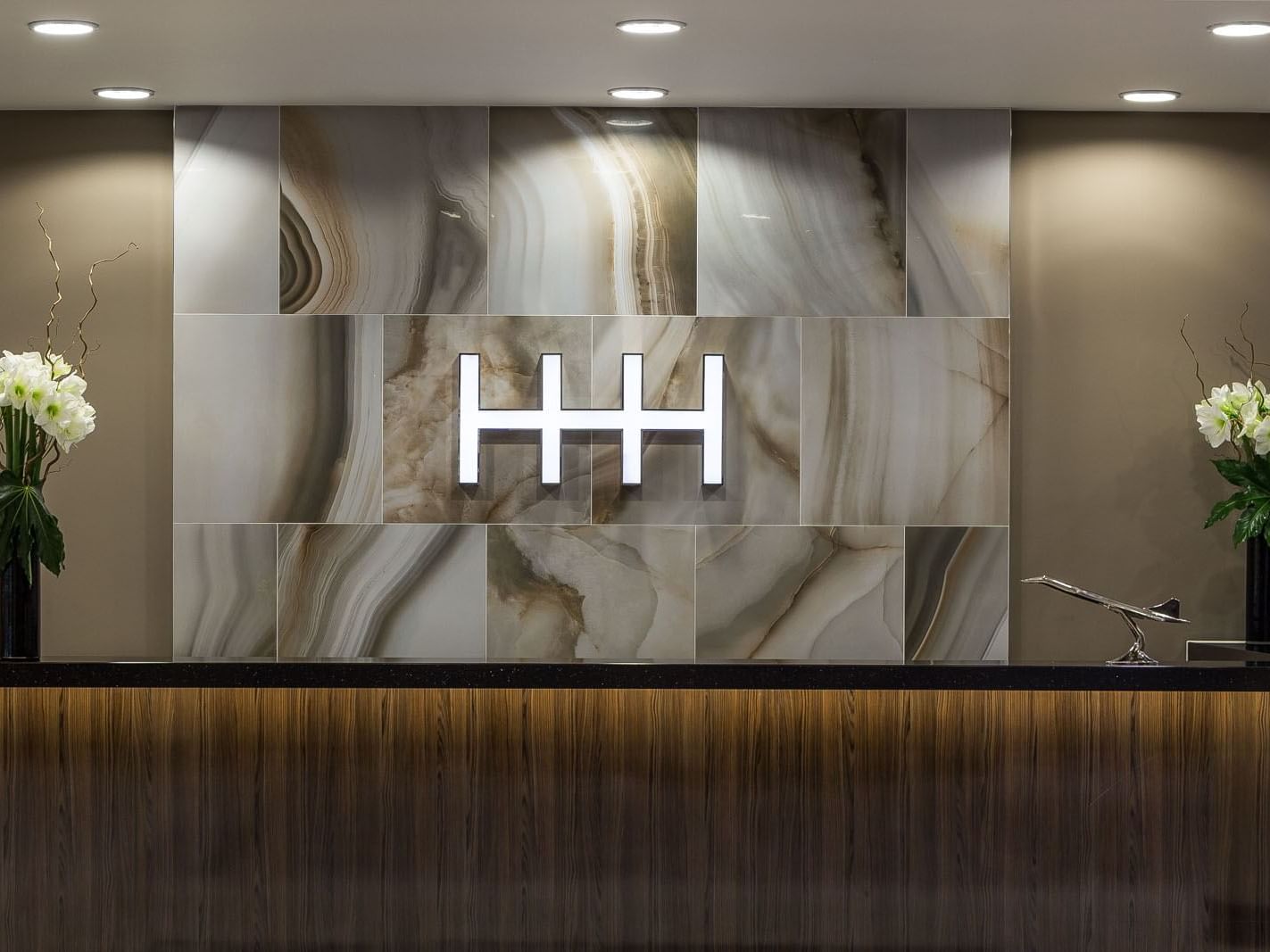 Reception desk with wooden front, marble wall backdrop, glowing logo, and flower arrangements at Heston Hyde Hotel