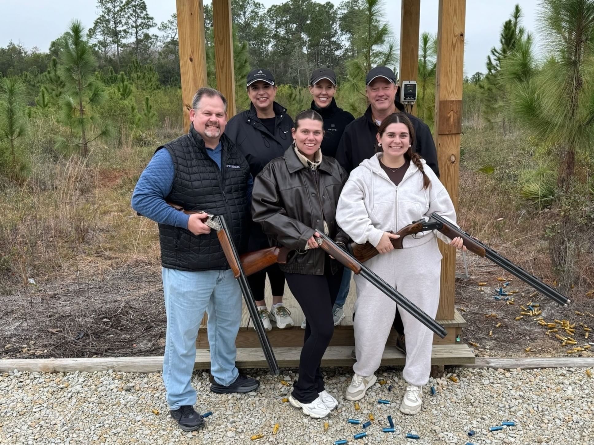 Group of six people posing with shotguns, standing by a wooden structure in a wooded area.