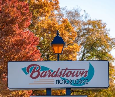 Retro-style sign with lit lamp post displaying dining hours against vibrant autumn foliage at Bardstown Motor Lodge