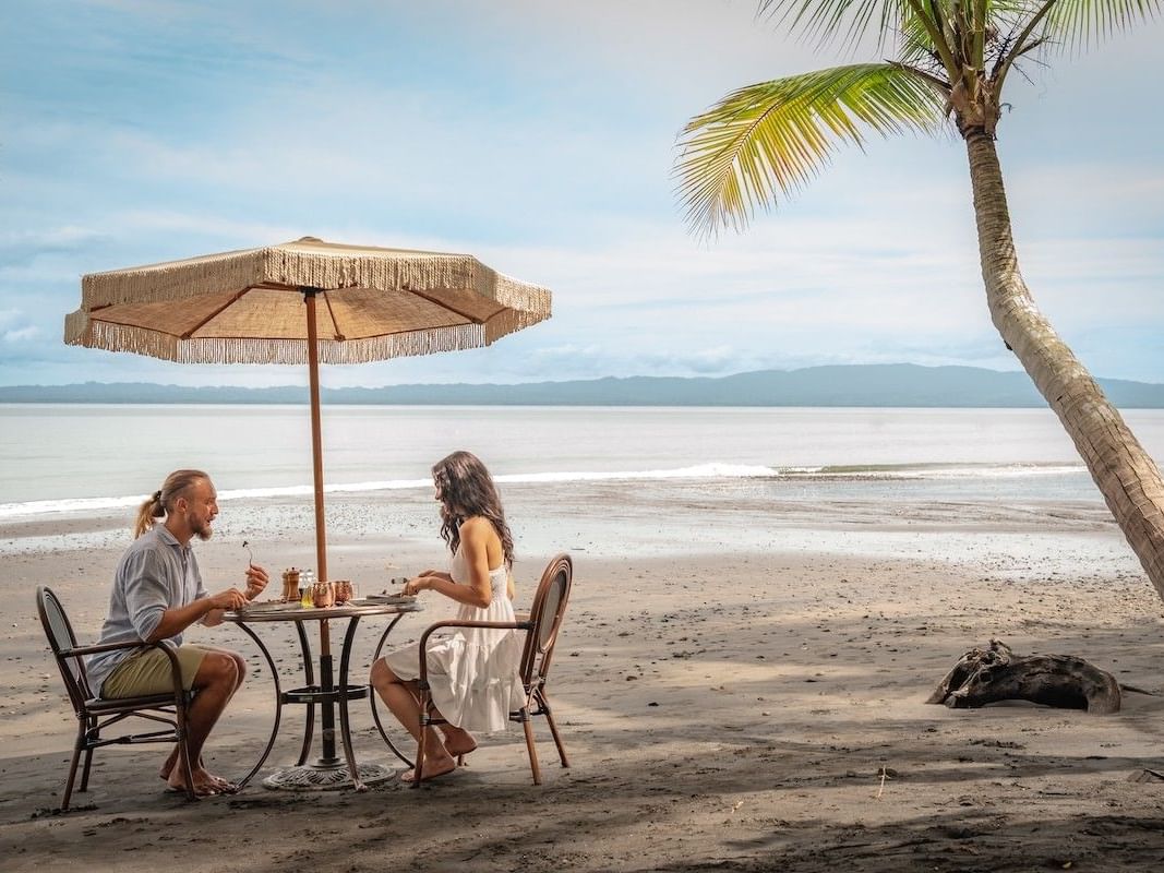 Couple enjoying a Castaway Beach Picnic under a fringed umbrella by the ocean at Playa Cativo Lodge in Golfito