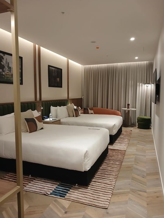 Two beds with patterned rugs by a curved sofa under recessed lighting in the guest room at the stylish Amora Hotel Group