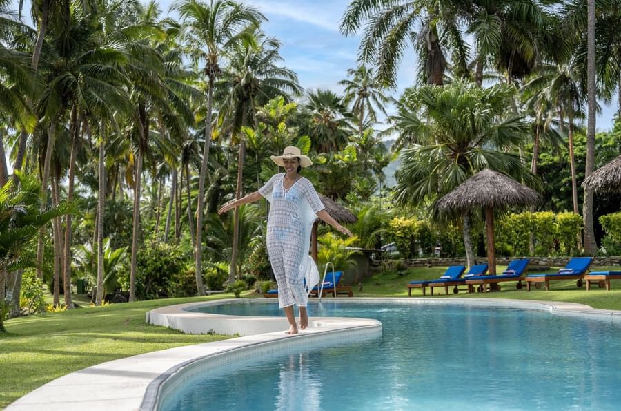 Woman in a white cover-up walking by a curved pool under tropical palm trees at Tambua Sands Beach Resort