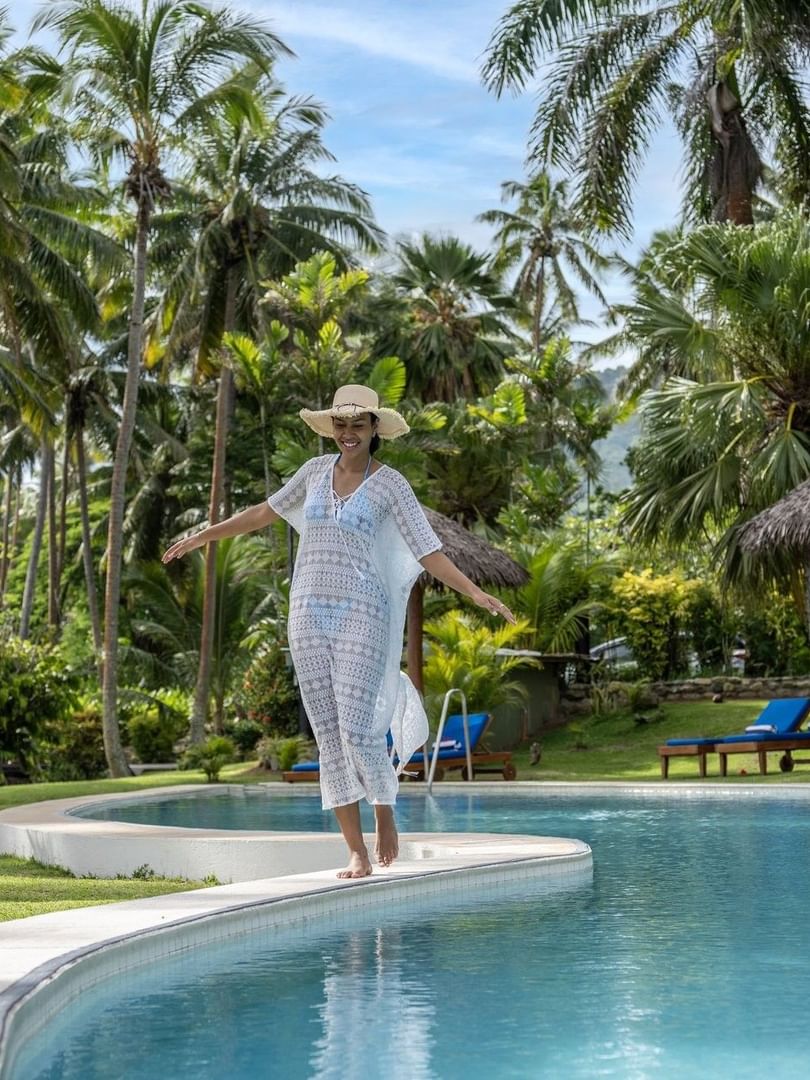 Woman in a white cover-up walking by a curved pool under tropical palm trees at Tambua Sands Beach Resort