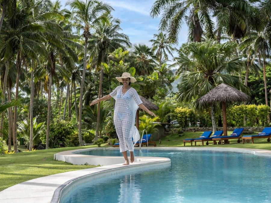 Woman in a white cover-up walking by a curved pool under tropical palm trees at Tambua Sands Beach Resort
