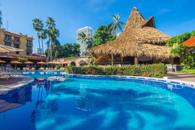 Large outdoor pool with lounge chairs, palm trees, and thatched roof hut in a tropical resort setting.