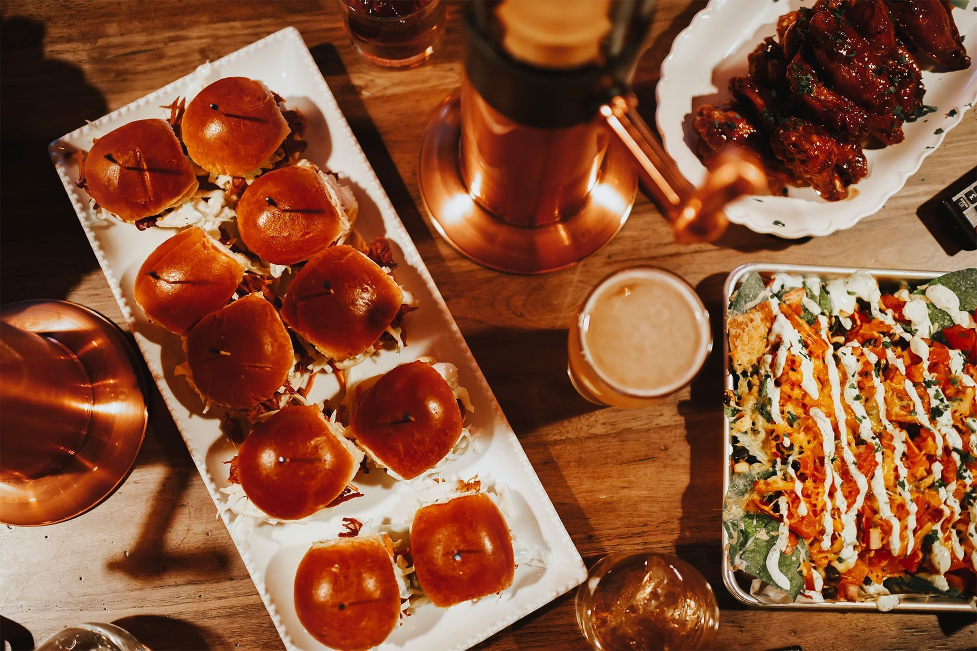 Barbecue sliders, sauced chicken wings, loaded nachos, and drinks arranged across a wooden dining table at The Brook