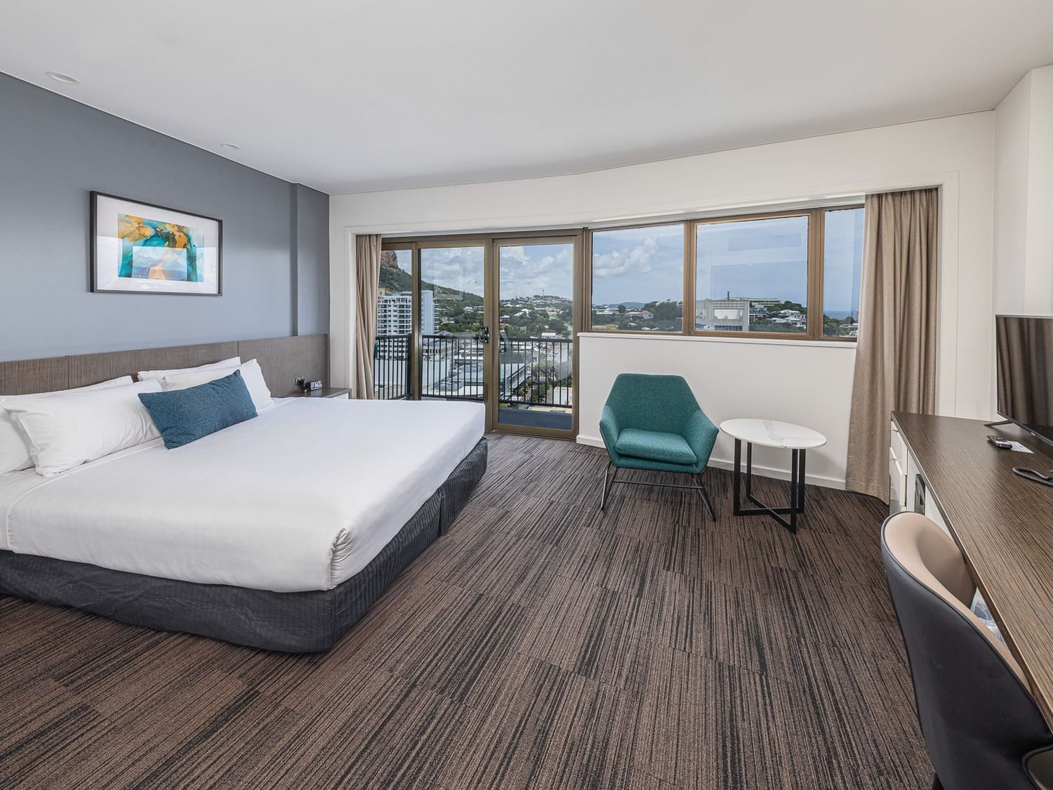 Image Details Alt Text: A bright, modern hotel room featuring a large bed with crisp white linens and a blue accent pillow against a dusty blue feature wall. The room includes a long wooden workspace with a flat-screen TV, a teal armchair with a small mar
