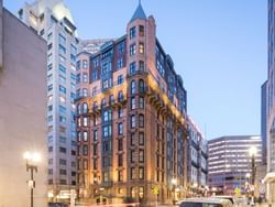 Dusk view of a city street with a prominent, illuminated, historic building among other structures.
