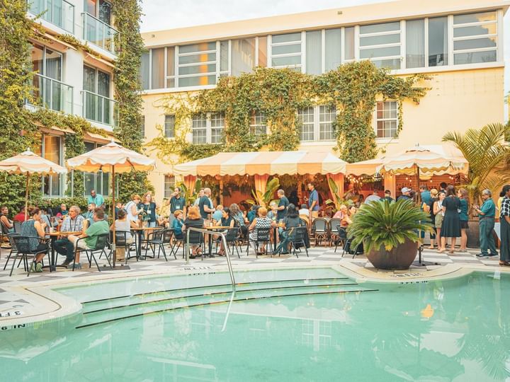 A poolside scene with people sitting at tables under umbrellas, surrounded by a building with ivy and palm trees.