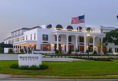 Exterior view of The White House Hotel, with white columns and an American flag on a sunny day