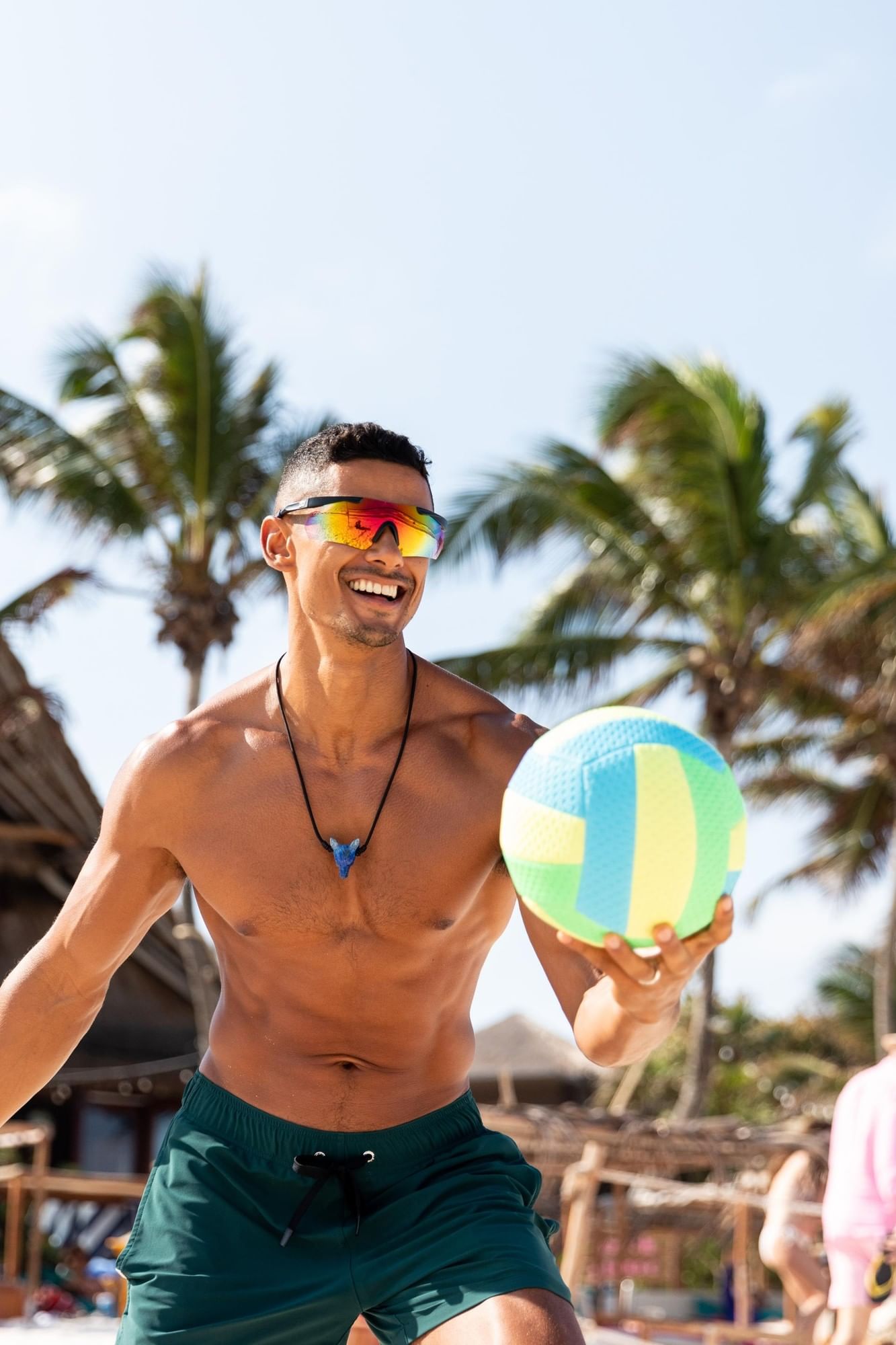 A smiling man in sunglasses holds a bright beach ball on the sandy shores near La Zebra Hotel.