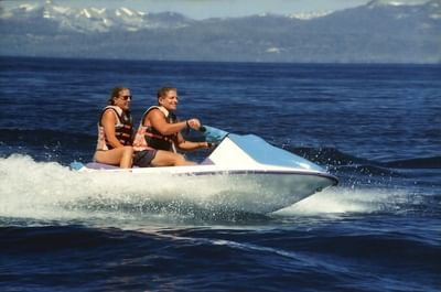 A couple enjoys riding a jet ski on the waters of Lake Tahoe with snow-capped mountains visible near Americana Village Suites