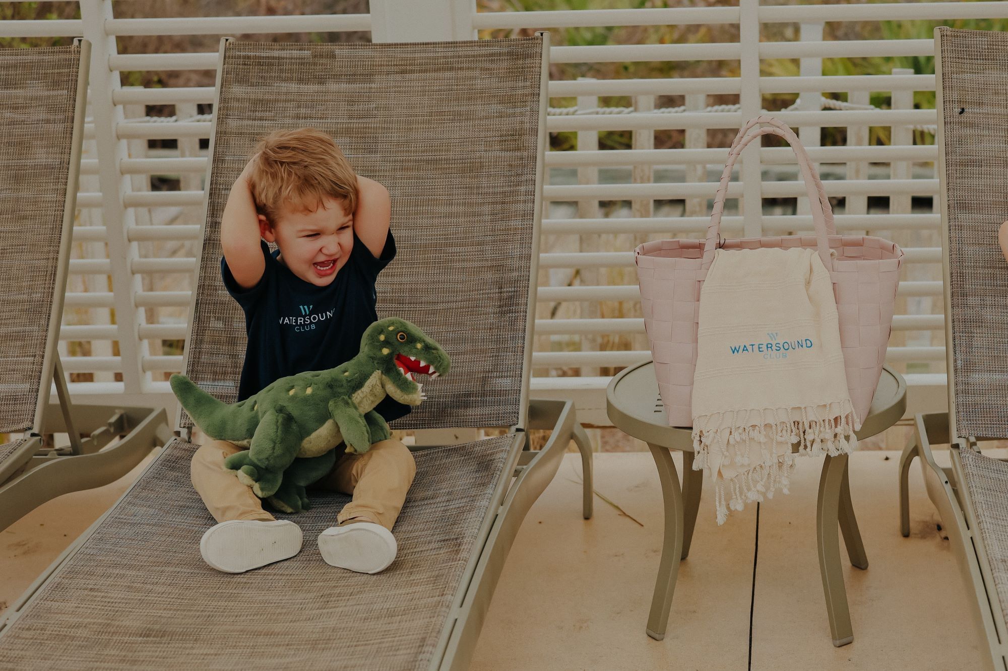 A young boy with dinosaur toy on lounge chair next to a table with pink bag and towel.