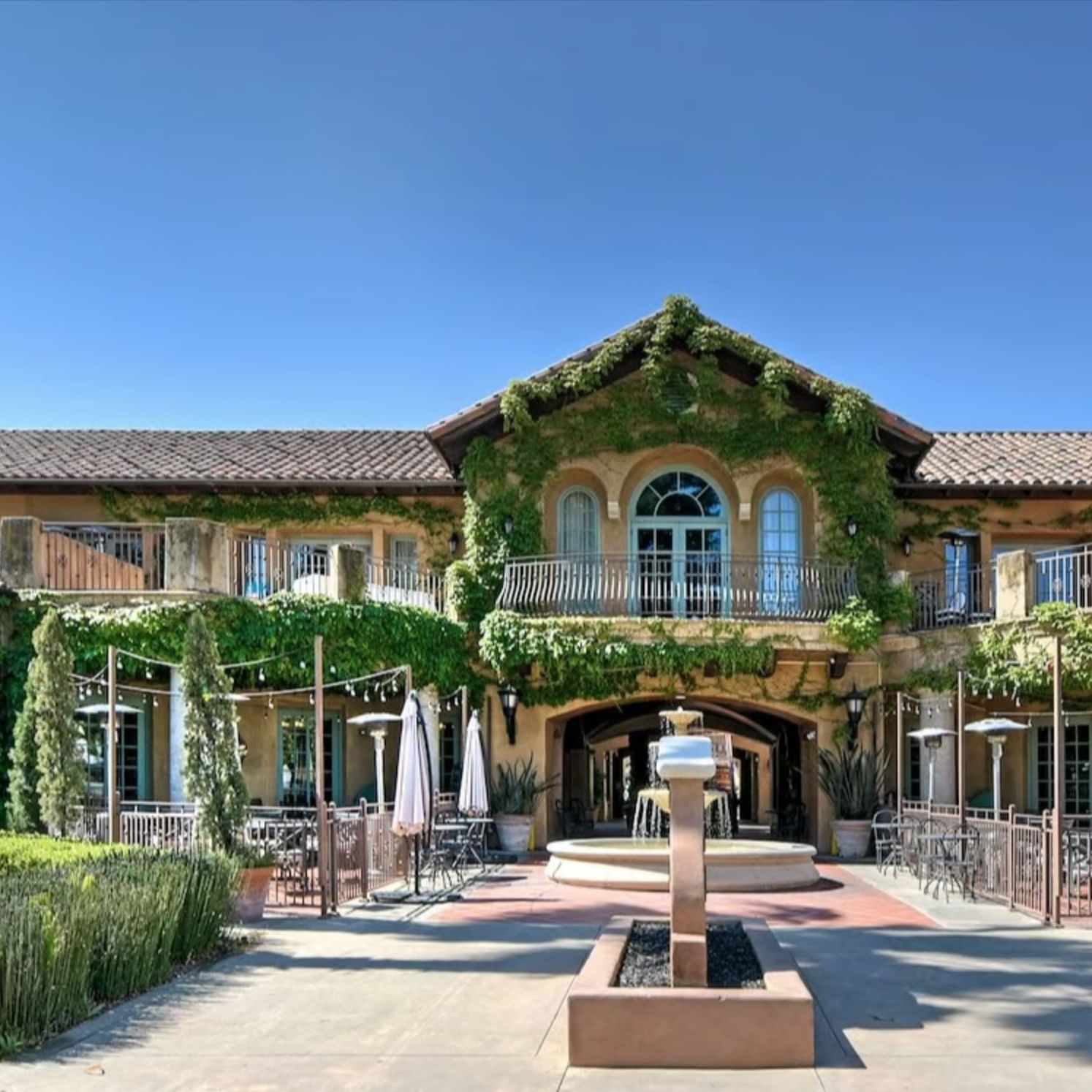 Front of a building with greenery, a fountain, and outdoor seating area with tables and umbrellas.