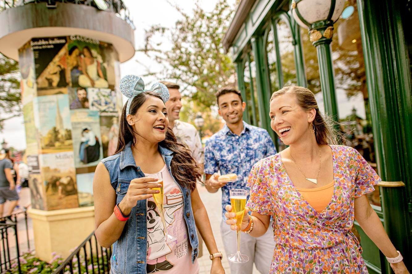 Two women holding Champagne flutes walk in front of two men with food in an outdoor setting.