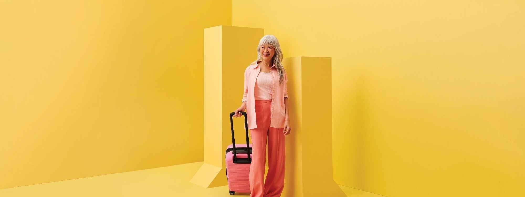 Woman in pink outfit with rolling suitcase standing against vibrant yellow geometric backdrop for stay, sip and shop