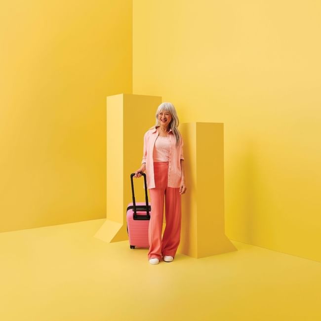 Woman in pink outfit with rolling suitcase standing against vibrant yellow geometric backdrop for stay, sip and shop