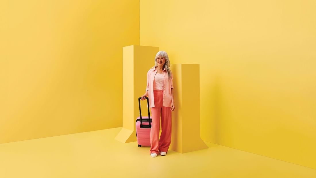 Woman in pink outfit with rolling suitcase standing against vibrant yellow geometric backdrop for stay, sip and shop