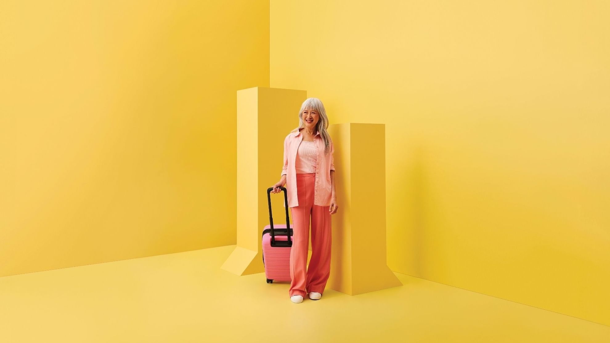Woman in pink outfit with rolling suitcase standing against vibrant yellow geometric backdrop for stay, sip and shop