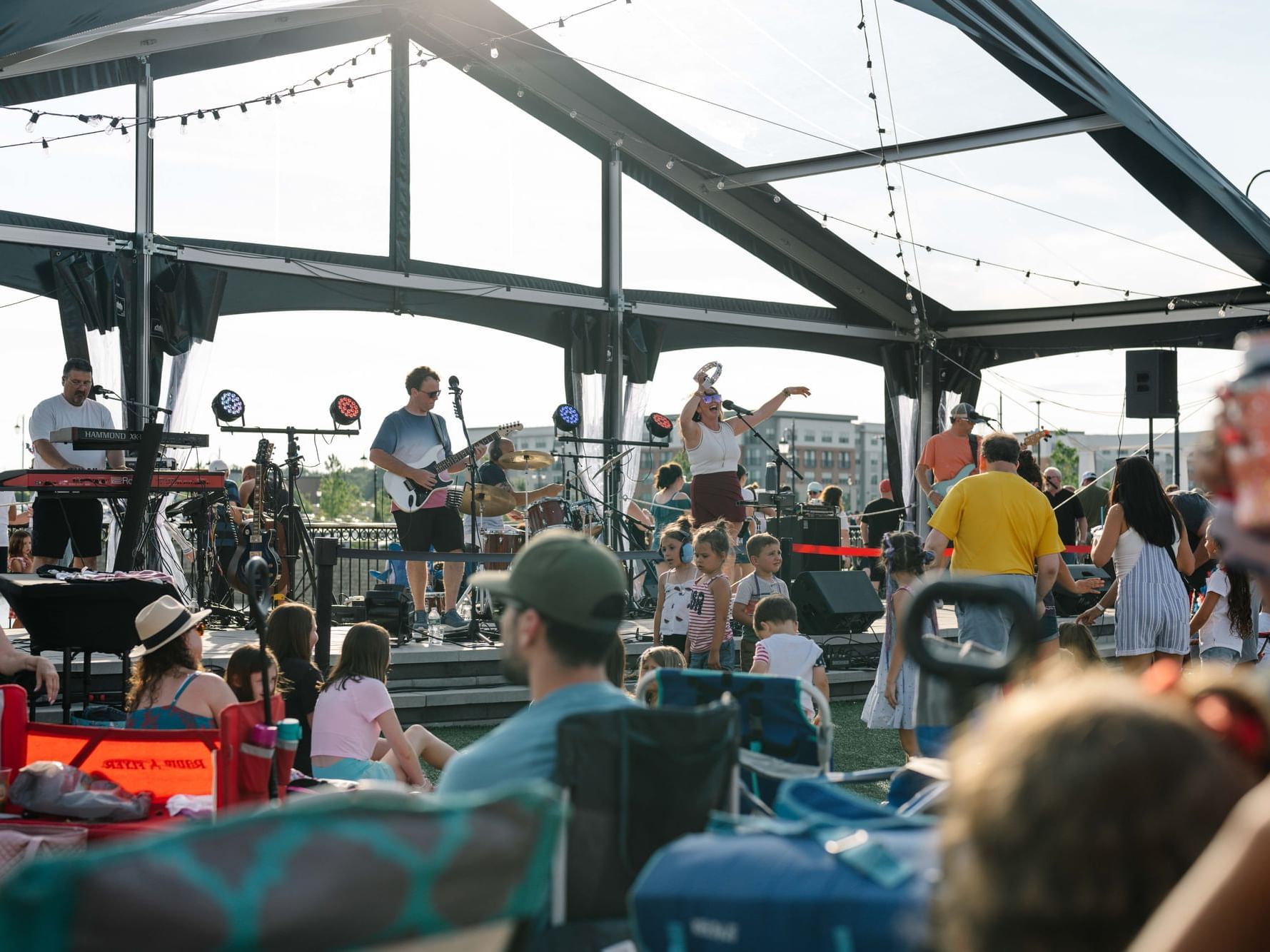 Outdoor concert under a canopy with a band performing on stage and audience seated in lawn chairs.