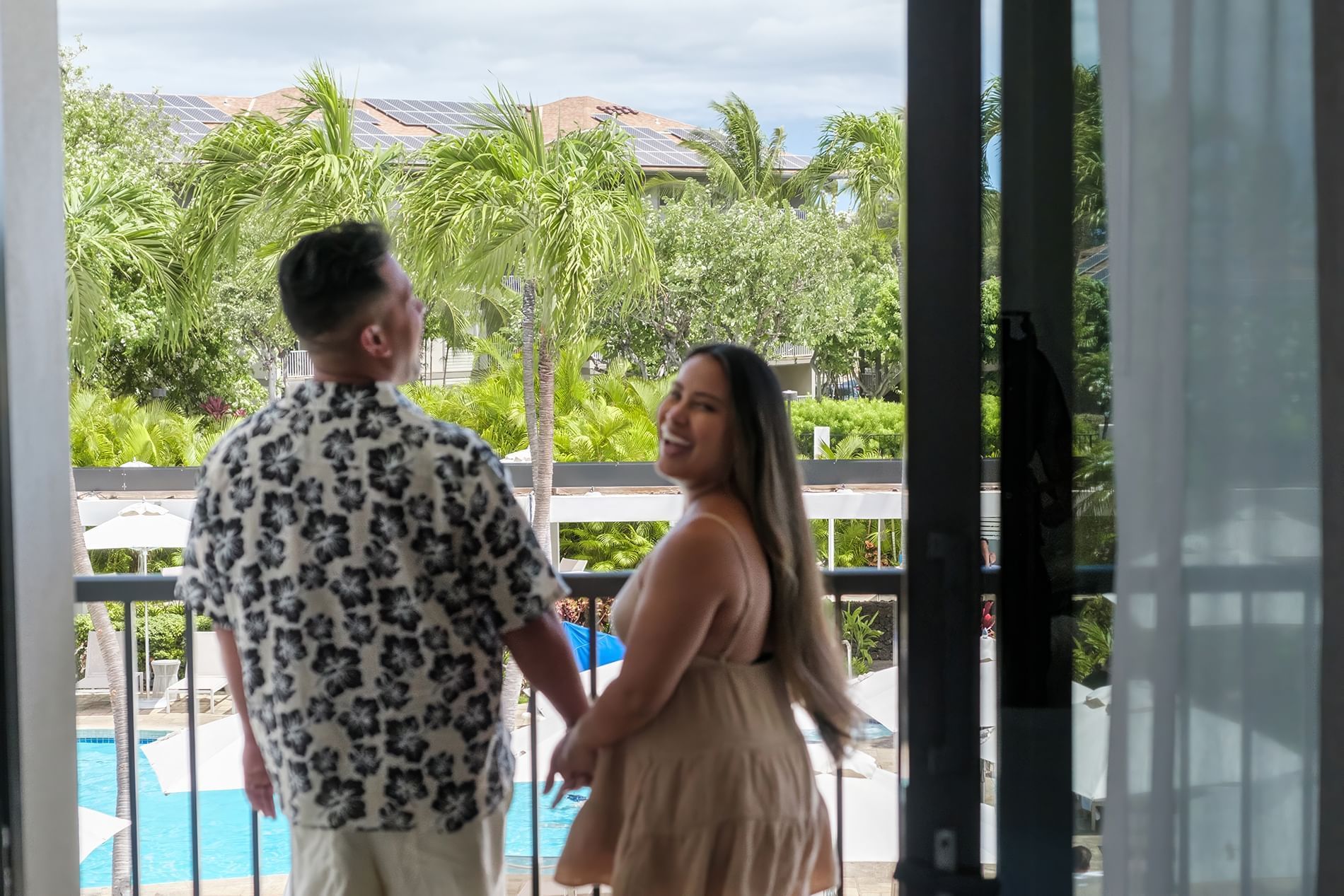 A couple stands on a balcony overlooking a pool and palm trees.