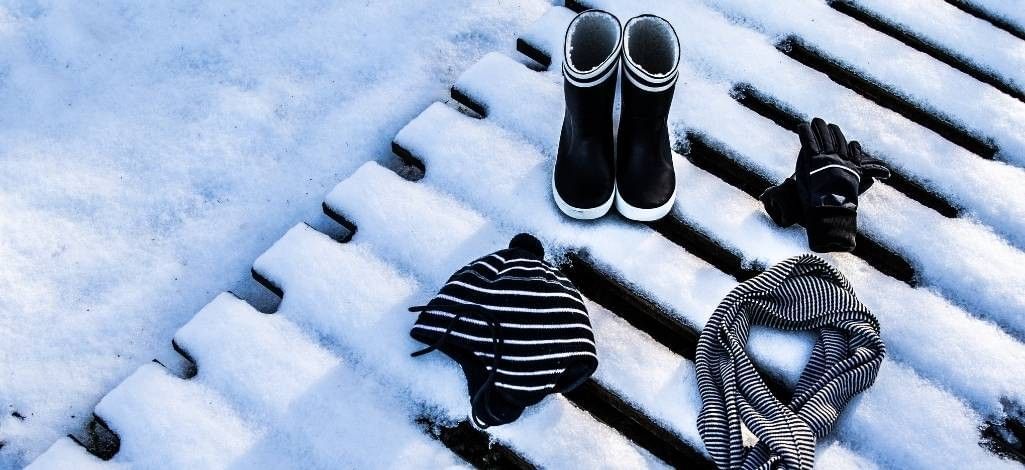 Winter clothing items—a pair of black boots, gloves, a striped hat and a striped scarf—laid out on a snow-covered wooden surface.