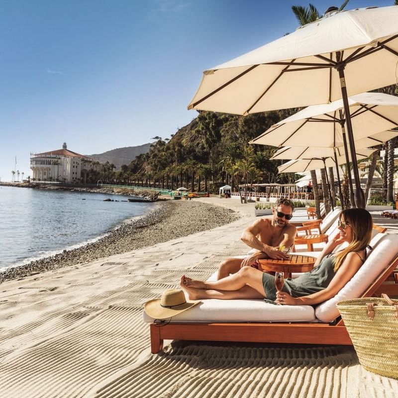 Two people relaxing on lounge chairs under umbrellas on a beach with a mountain in the background.