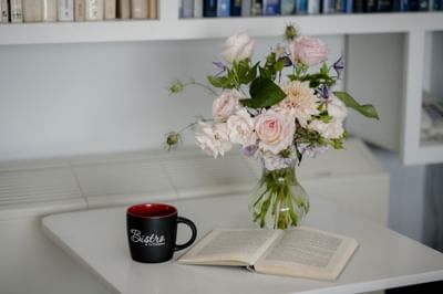 Black coffee mug with open book and fresh flower vase on a white table at La Tourelle Hotel and Spa