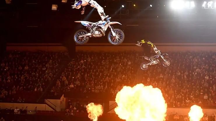 Freestyle motocross rider mid-air during a backflip stunt with fireworks and lights in the background.