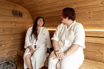Two happy women in white robes relaxing in a wood barrel sauna during fun things to do in Moncton at Rodd Moncton