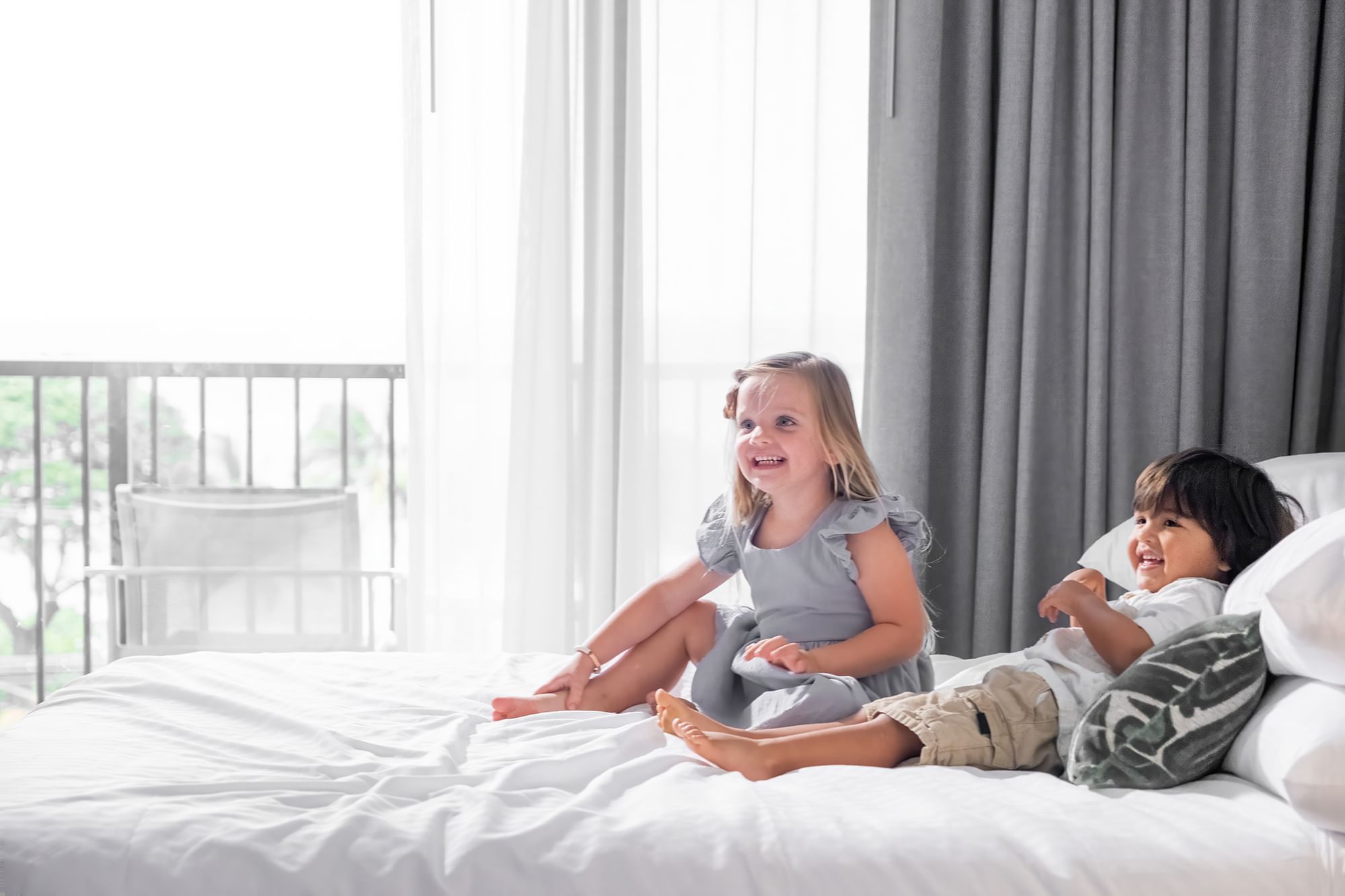 Two children playing on a bed in a hotel room with a large window and balcony.