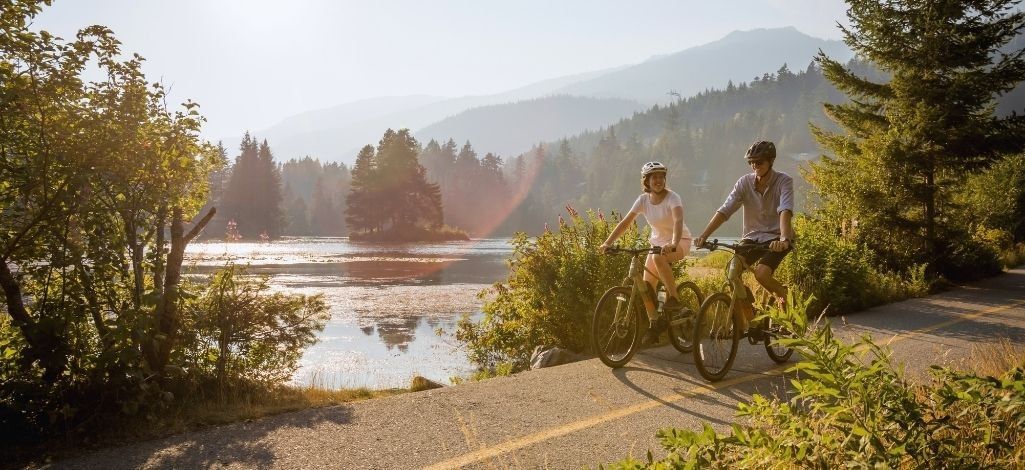 Cyclists ride beside a quiet Whistler lake on a paved trail surrounded by forest and mountain views