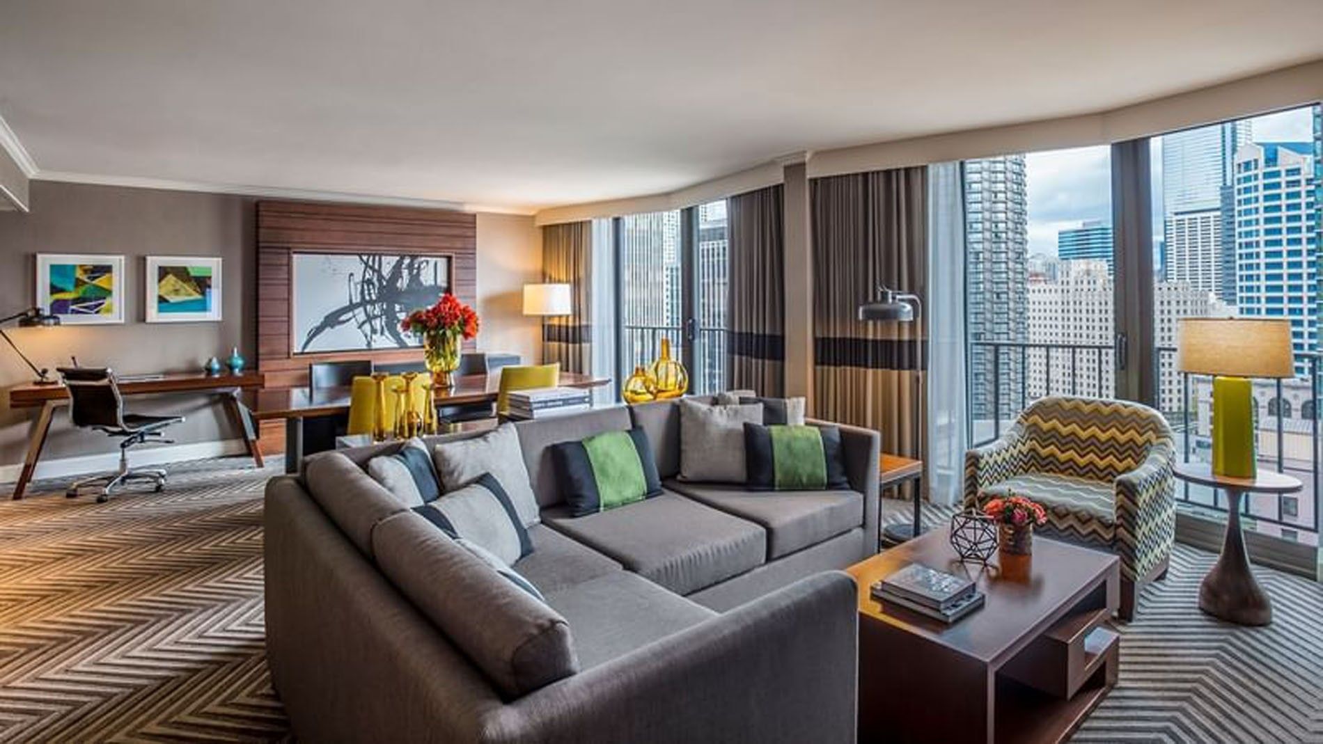 A wide view of a hotel lobby with a sofa, coffee table, desk, chair, and city view.