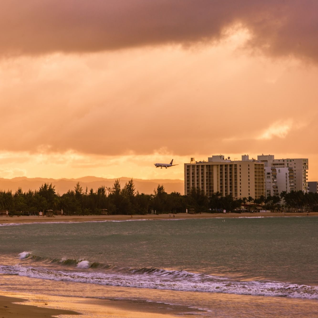 Airplane flying over the beach with buildings in the background at sunset.