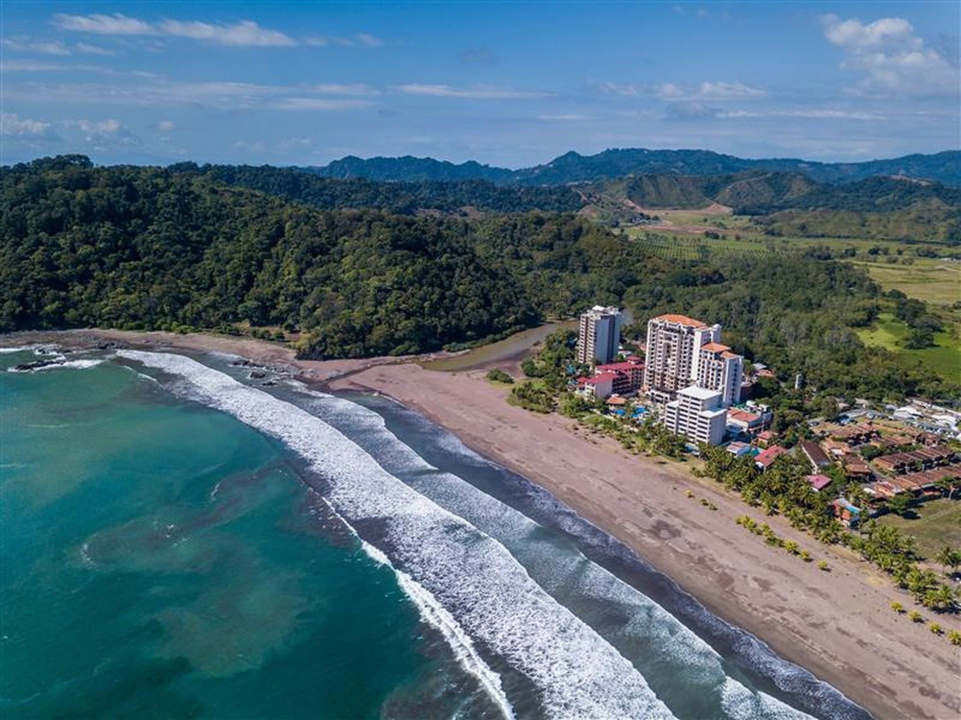 Aerial view of dark sand beach and turquoise waves by coastline, with a resort on the edge near Santa Lucia Jungle Hacienda