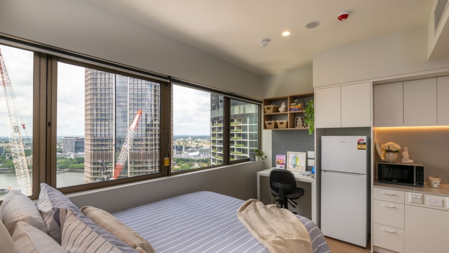 Modern student accom room with bed, desk, refrigerator, and city view through large windows.