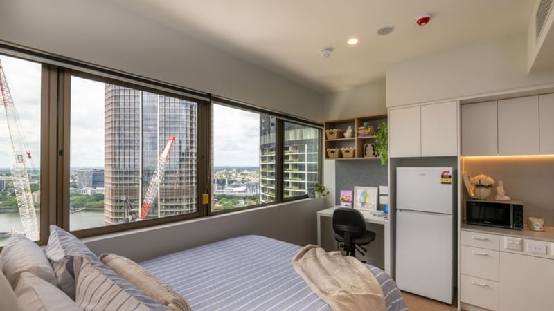 Modern student accom room with bed, desk, refrigerator, and city view through large windows.