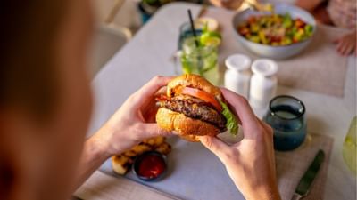 Person holding a large juicy beef burger with tomato and lettuce over a dining table at Sugar Cane Club Hotel & Spa