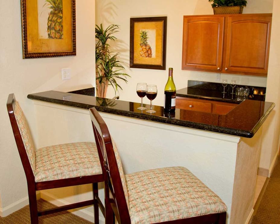 A kitchenette with two bar stools, a counter with wine glasses and a bottle, and wooden cabinets.
