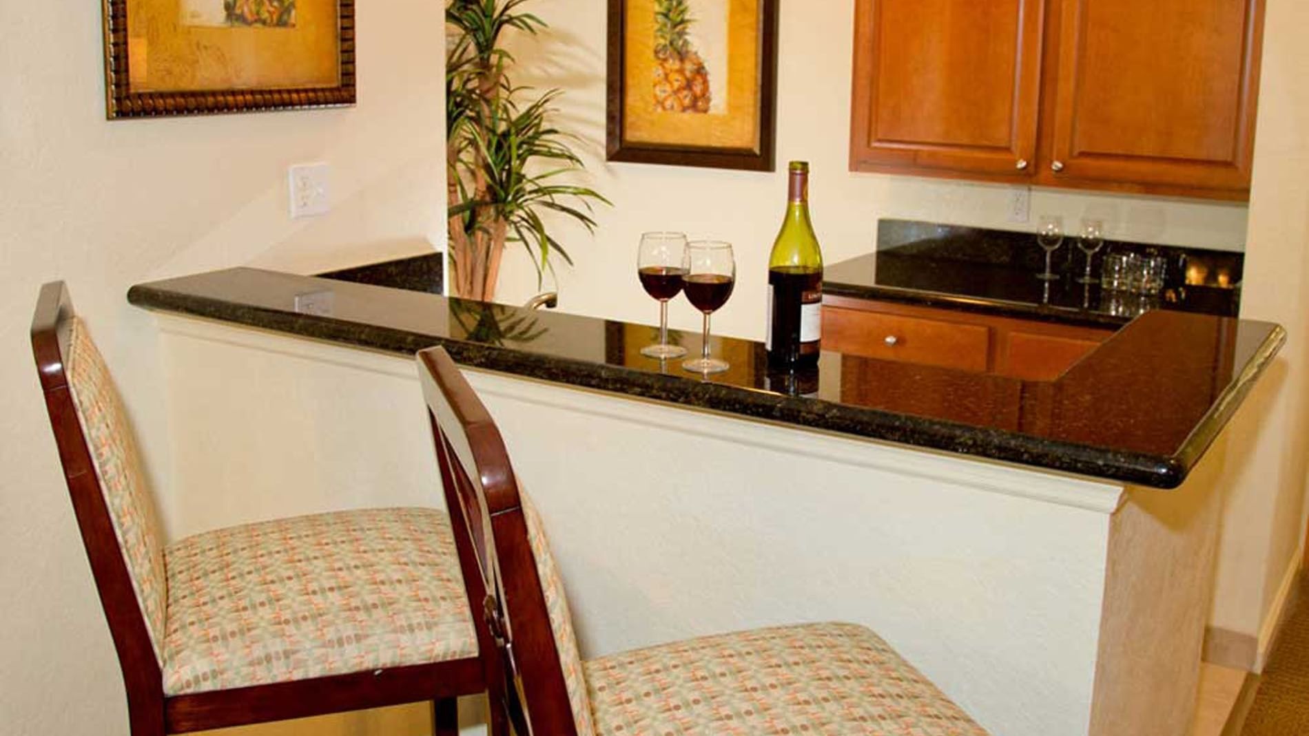 A kitchenette with a black granite countertop, two bar stools, a bottle of wine, and two glasses.