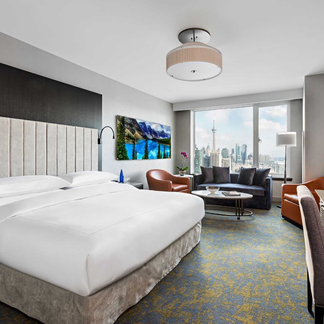 A hotel room with a large bed, couch, table, and cityscape view through large windows.