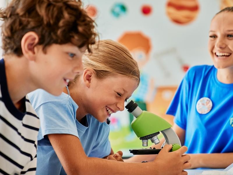 Tre bambini guardano attraverso un microscopio verde in una stanza colorata.
