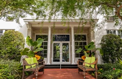 Elegant brick and white column entrance of the Tradewinds Apartment Hotel building framed by trees and wooden benches