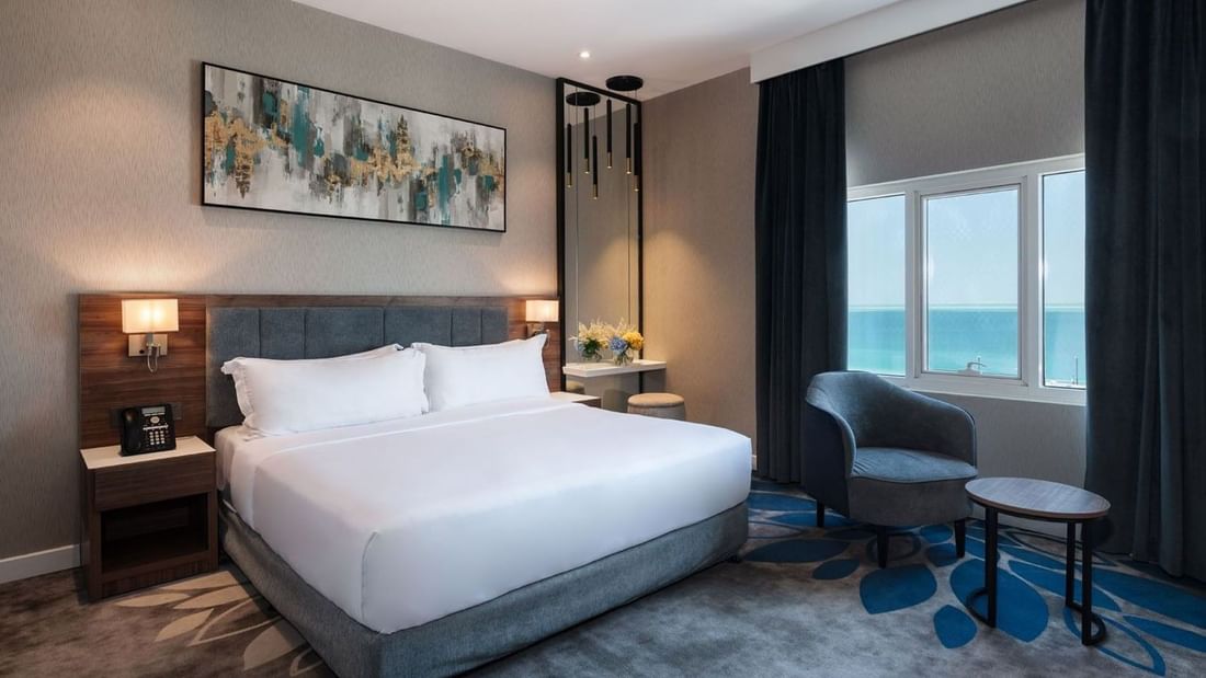 White bed by a dark armchair near a large window in Executive Room Sea View at Warwick Al Jubail