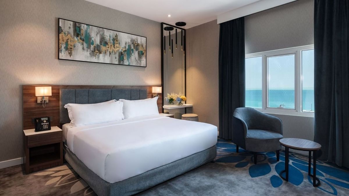 White bed by a dark armchair near a large window in Executive Room Sea View at Warwick Al Jubail