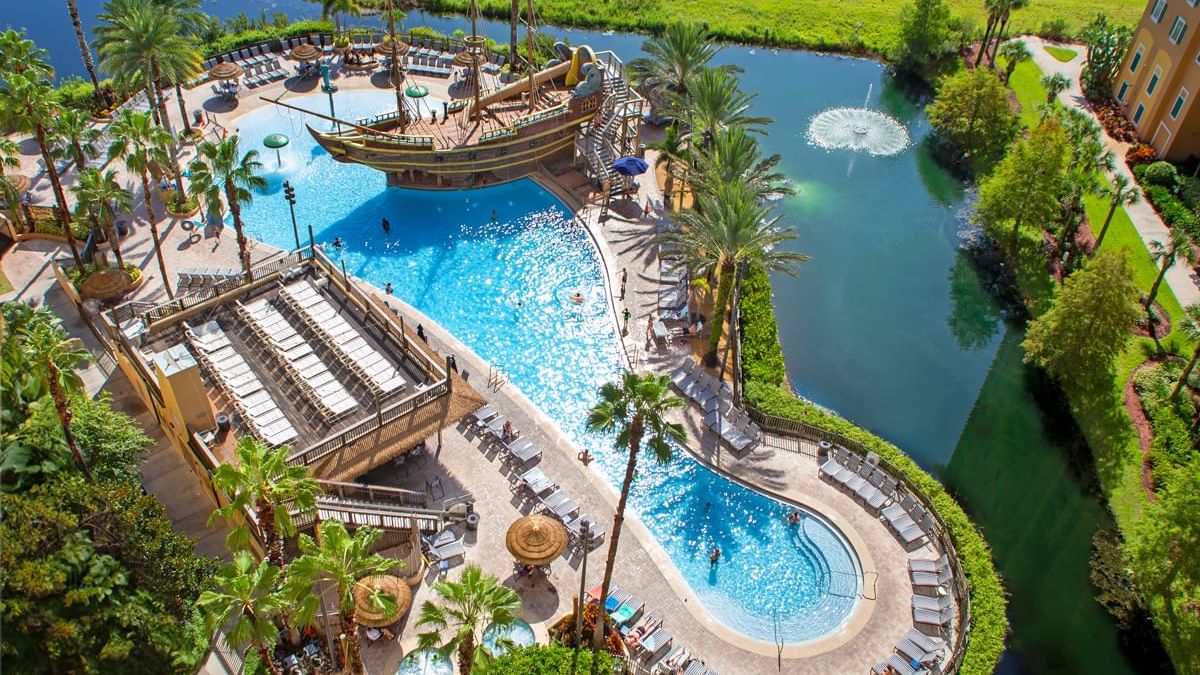 Aerial view of a large pool area with a pirate ship playground, lounge chairs, and palm trees.