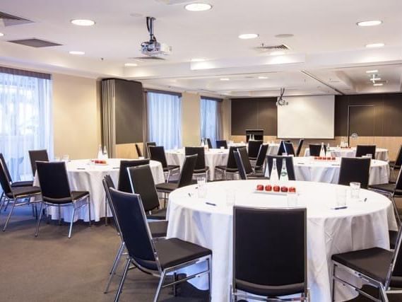 Bright conference room set with multiple round tables, black chairs, and a large projector screen at The Sebel Brisbane